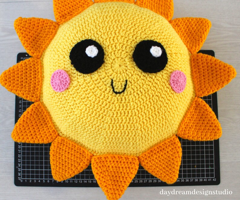 May include: A crocheted sun pillow with a yellow face, orange rays, and black eyes. The sun has pink cheeks and a smiling mouth.