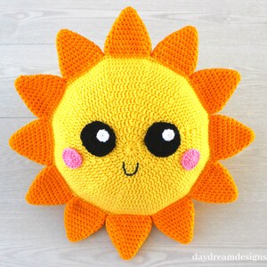 May include: A crocheted sun pillow with a yellow face, orange rays, black eyes, white pupils, and pink cheeks. The sun is smiling.
