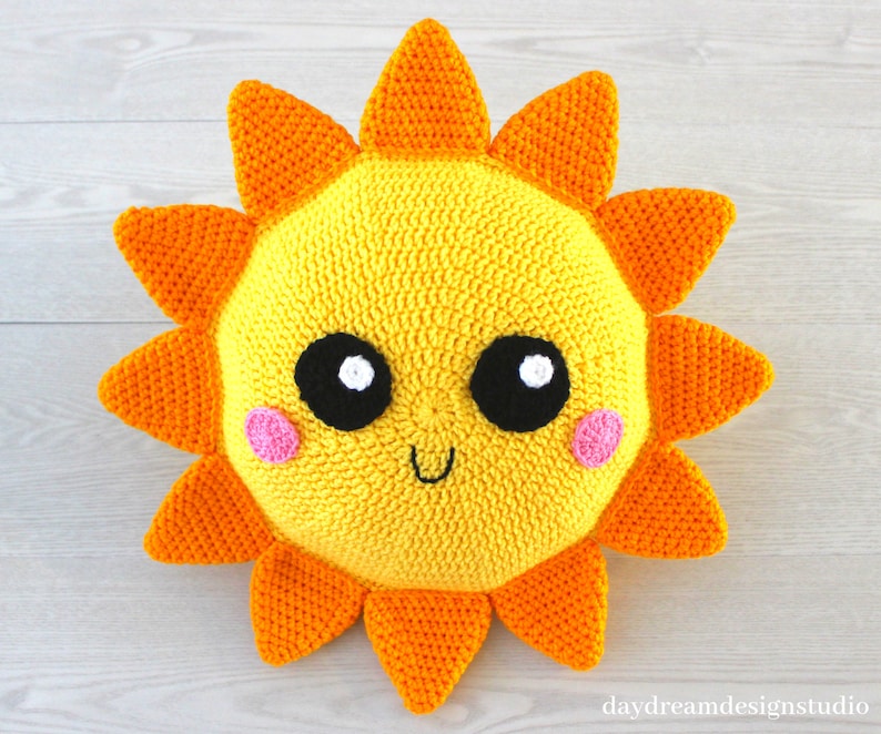 May include: A crocheted sun pillow with a yellow face, orange rays, black eyes, white pupils, and pink cheeks. The sun is smiling.