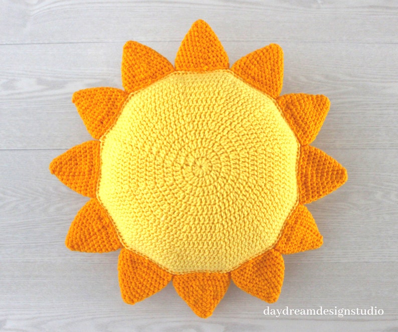 May include: A crocheted sun pillow with yellow and orange yarn. The pillow is shaped like a sun with a round center and 12 points.