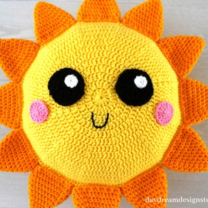 May include: A crocheted sun pillow with a yellow face, orange rays, and pink cheeks. The sun has large black eyes with white highlights and a smiling mouth.