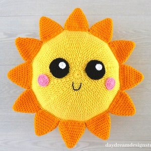 May include: A crocheted sun pillow with a yellow face, black eyes, white highlights, pink cheeks, and a smiling mouth. The sun has orange rays.