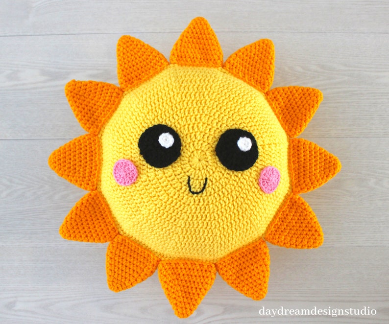 May include: A crocheted sun pillow with a yellow face, black eyes, white highlights, pink cheeks, and a smiling mouth. The sun has orange rays.