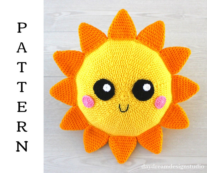 May include: A crocheted sun pillow with a yellow face, orange rays, black eyes, white pupils, and pink cheeks. The sun is smiling.