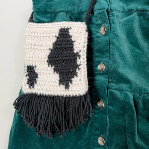 May include: A small crocheted bag with black and white cow print design and black fringe hanging from a green corduroy dress.