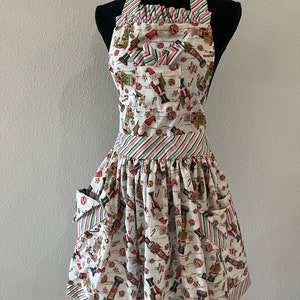 May include: A white apron with a red and green striped trim. The apron has a festive print of nutcrackers, candy canes, and ornaments. It has a large pocket on the front.