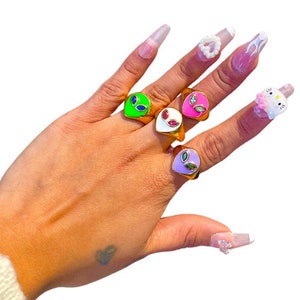 May include: Five gold rings with different colored enamel alien faces. The rings are on a hand with painted nails.