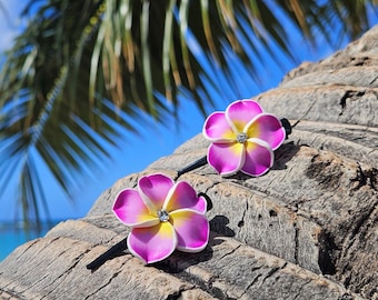May include: Two pink and yellow flower hair clips with a rhinestone center. The clips are on a brown wooden surface.