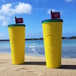 May include: Two yellow reusable Starbucks cups with a textured surface and green lids. The cups have a small red flag with the text "Hawaii Collection" on top. The cups are on a sandy beach with a blue ocean and sky in the background.