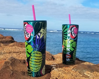 May include: Two green stainless steel tumblers with colorful tropical fruit and leaf designs. The tumblers have pink straws and the text "HAWAII COLLECTION" and "STARBUCKS" printed on them. The tumblers are sitting on a rocky surface with a blue ocean in the background.