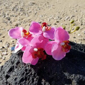 May include: A cluster of pink orchid flowers with red centers, arranged on a dark gray rock. The flowers are attached to a silver clip.