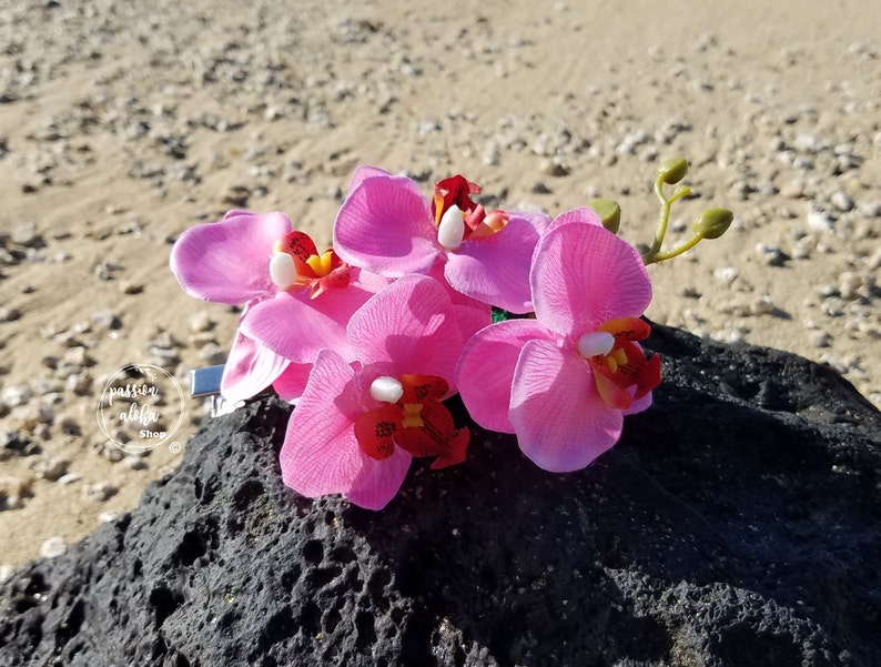 May include: A cluster of pink orchid flowers with red centers, arranged on a dark gray rock. The flowers are attached to a silver clip.