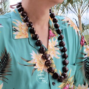 May include: A brown seed lei necklace worn by a person wearing a teal blue Hawaiian shirt with a floral print.