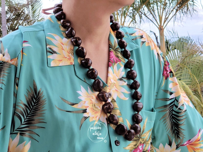 May include: A brown seed lei necklace worn by a person wearing a teal blue Hawaiian shirt with a floral print.