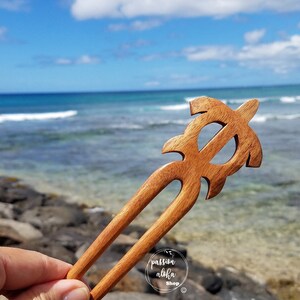 May include: Wooden hair stick with a carved turtle design. The stick is brown and has a natural finish. The background is a blurry image of the sea.