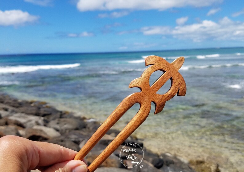 May include: Wooden hair stick with a carved turtle design. The stick is brown and has a natural finish. The background is a blurry image of the sea.