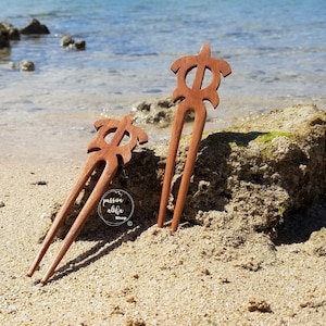 May include: Two wooden hair forks with a turtle design. The forks are brown and have a natural wood finish. The forks are on a sandy beach with a rock in the background.