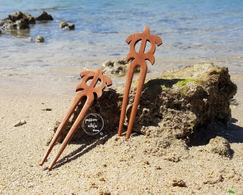 May include: Two wooden hair forks with a turtle design. The forks are brown and have a natural wood finish. The forks are on a sandy beach with a rock in the background.