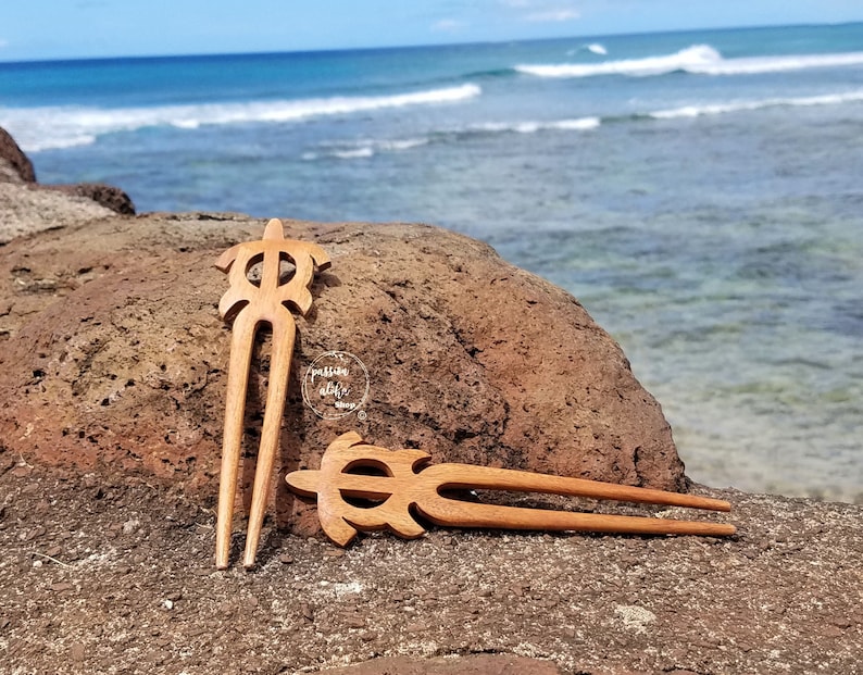 May include: Two wooden hair sticks with a turtle design. The sticks are brown and have a smooth finish. They are lying on a brown rock with a blurred background of the sea.