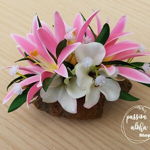May include: A pink and white flower arrangement with green leaves, set on a piece of brown wood. The flowers are arranged in a cluster and are surrounded by green leaves. The arrangement is sitting on a piece of brown wood. The text "passion aloha Shop" is visible in the lower right corner of the image.