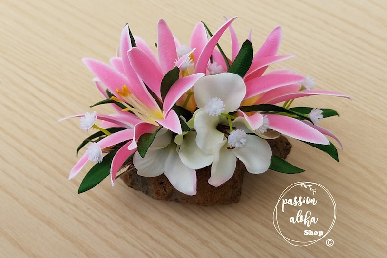 May include: A pink and white flower arrangement with green leaves, set on a piece of brown wood. The flowers are arranged in a cluster and are surrounded by green leaves. The arrangement is sitting on a piece of brown wood. The text "passion aloha Shop" is visible in the lower right corner of the image.