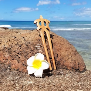 May include: A wooden hair fork shaped like a turtle, with a white plumeria flower on a brown rock, against a blue ocean background.