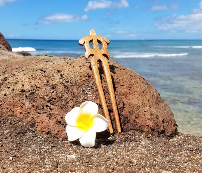 May include: A wooden hair fork shaped like a turtle, with a white plumeria flower on a brown rock, against a blue ocean background.