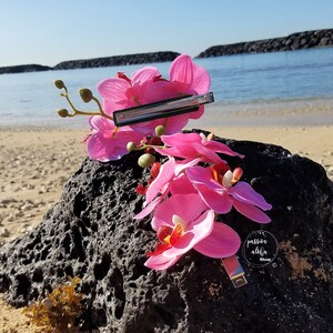 May include: Pink orchid hair clip with a silver barrette. The clip is on a black rock with a beach in the background.