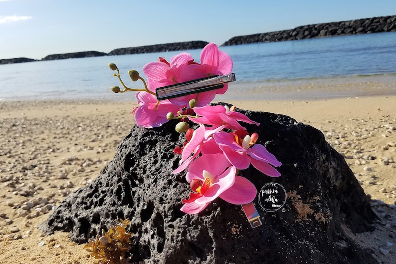 May include: Pink orchid hair clip with a silver barrette. The clip is on a black rock with a beach in the background.