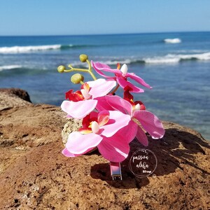 May include: Pink orchid flower hair clip with a silver metal bar. The clip is on a brown rock with a blue ocean and white waves in the background. The text "passion aloha shop" is on the clip.