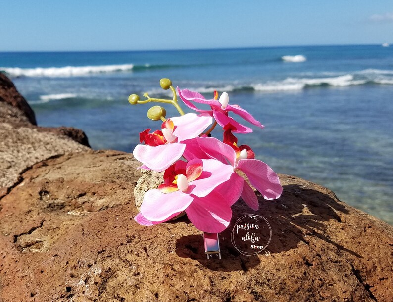 May include: Pink orchid flower hair clip with a silver metal bar. The clip is on a brown rock with a blue ocean and white waves in the background. The text "passion aloha shop" is on the clip.