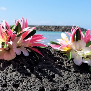 May include: Two pink and white flower hair clips with green leaves, set against a backdrop of a rocky beach and blue ocean.