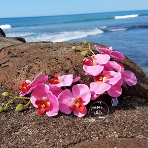 May include: Pink orchid flower hair clip with a silver bar clip. The flowers are arranged in a circle and are on a brown rock with a blue ocean in the background. The text "passion aloha shop" is visible in the image.