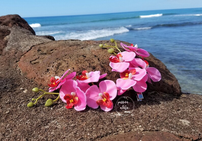 May include: Pink orchid flower hair clip with a silver bar clip. The flowers are arranged in a circle and are on a brown rock with a blue ocean in the background. The text "passion aloha shop" is visible in the image.