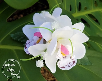 May include: White orchid flower hair clip with pink accents and white and purple speckles. The flower is on a green leaf background.