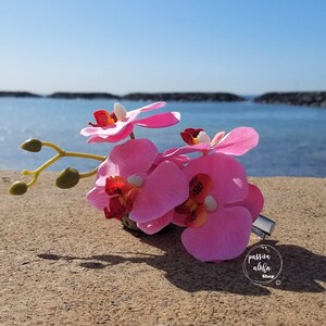 May include: Pink orchid flower hair clip with a silver metal clip. The clip is on a sandy beach with a blue ocean in the background.