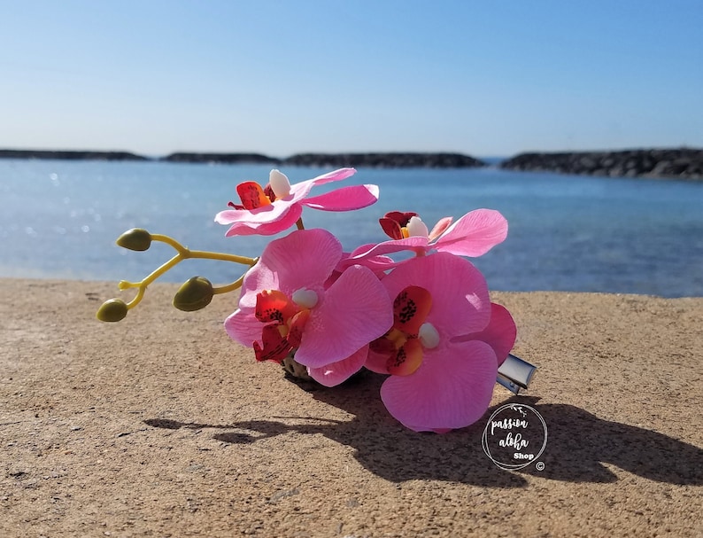 May include: Pink orchid flower hair clip with a silver metal clip. The clip is on a sandy beach with a blue ocean in the background.