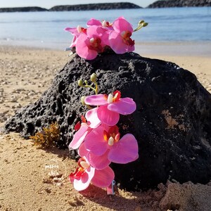 May include: Two pink orchid hair clips on a black rock on a sandy beach. The orchids are made of fabric and have a realistic appearance.