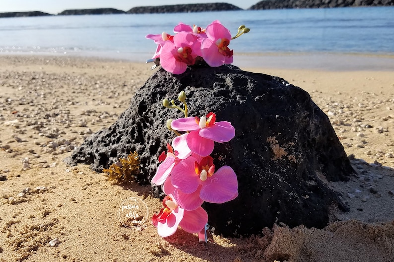 May include: Two pink orchid hair clips on a black rock on a sandy beach. The orchids are made of fabric and have a realistic appearance.