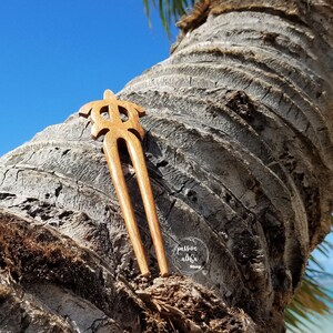 May include: A wooden hair fork with a turtle design. The fork is brown and has two prongs. It is resting on a tree trunk.