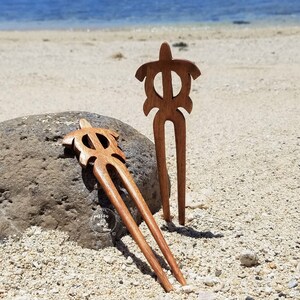 May include: Two wooden hair sticks with turtle designs. The sticks are brown and have a natural finish. They are lying on a sandy beach.