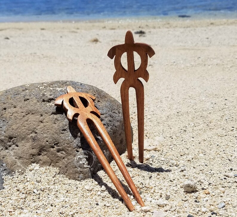 May include: Two wooden hair sticks with turtle designs. The sticks are brown and have a natural finish. They are lying on a sandy beach.