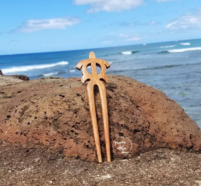 May include: Wooden hair fork with a turtle design. The fork is brown and has a natural finish. It is sitting on a rock with the ocean in the background. The text "passion aloha shop" is visible in the lower right corner of the image.