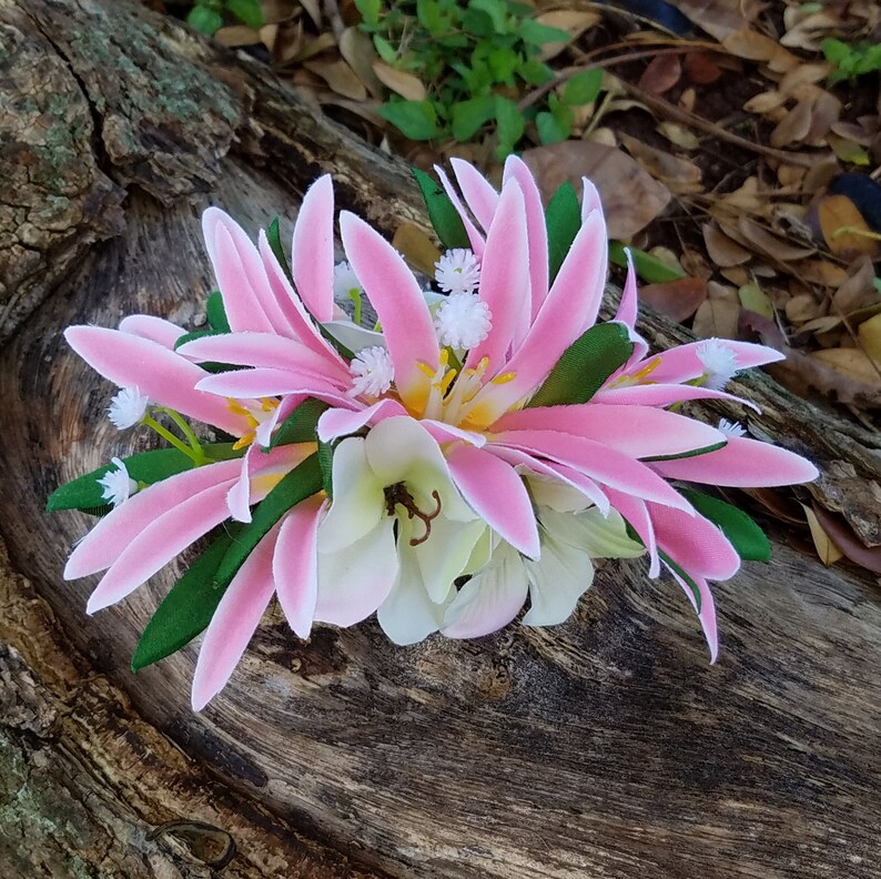 May include: A pink and white flower hair clip with green leaves. The flower is made of fabric and has a realistic appearance.