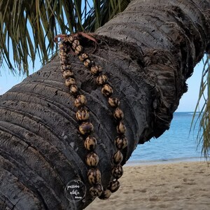 May include: A brown and black seed lei draped over a palm tree trunk with a beach and ocean in the background. The lei is made of natural materials and has a rustic look.