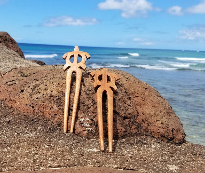 May include: Two wooden hair sticks featuring a turtle design. The sticks are brown and have a natural finish. They are resting on a rock with the ocean in the background.