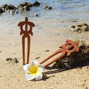 May include: Two wooden hair sticks with a turtle design. The sticks are brown and have a natural finish. The sticks are lying on a sandy beach with a white flower.