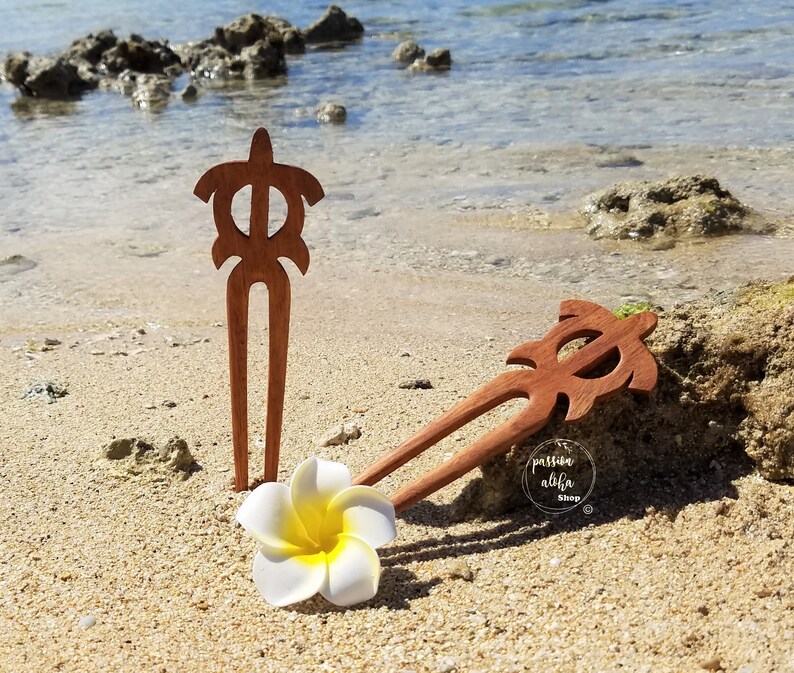May include: Two wooden hair sticks with a turtle design. The sticks are brown and have a natural finish. The sticks are lying on a sandy beach with a white flower.