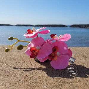 May include: Pink orchid flower hair clip with a green stem and leaves. The clip is on a sandy beach with a blue ocean in the background. The text "passion aloha shop" is visible in the lower right corner of the image.