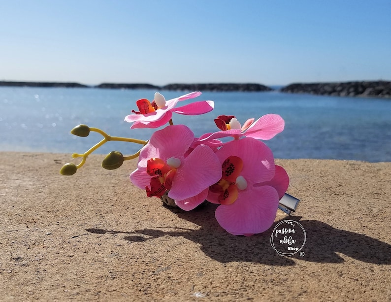 May include: Pink orchid flower hair clip with a green stem and leaves. The clip is on a sandy beach with a blue ocean in the background. The text "passion aloha shop" is visible in the lower right corner of the image.